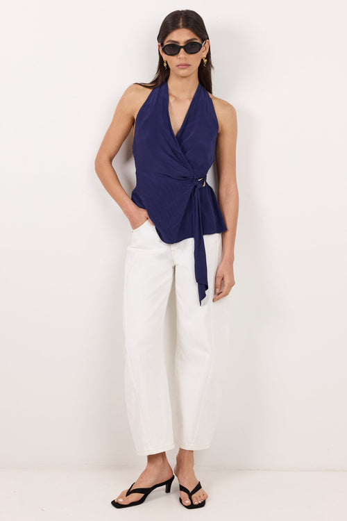 A model wearing a navy blue halter wrap top with a tie waist, white cropped pants, and black sandals, standing against a plain white studio background.