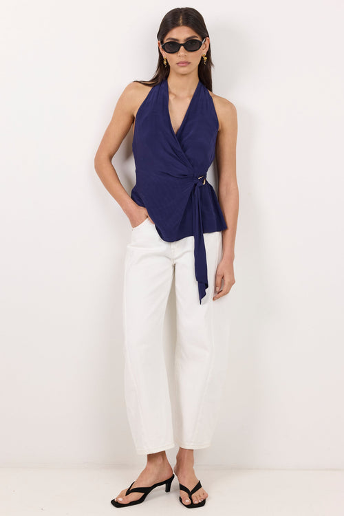 A woman modeling a navy blue halter wrap top with a tie waist, paired with white cropped pants and black sandals, standing against a plain white studio background.