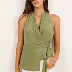 A woman modeling a sage green halter wrap top paired with white wide-leg pants and beige sandals, standing against a plain white studio background.