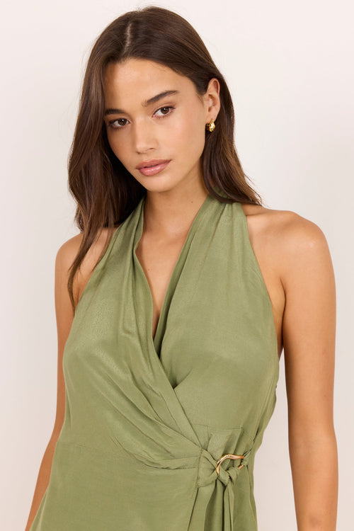 A woman wearing an olive green halter wrap dress with a twisted tie detail at the waist, photographed against a plain light background.