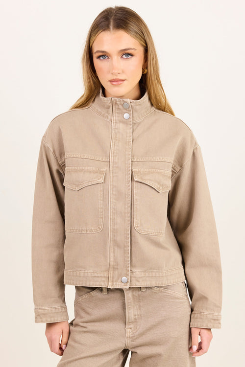 A woman modeling a cropped beige denim jacket with a stand-up collar and front flap pockets, paired with matching beige pants, against a plain light background.