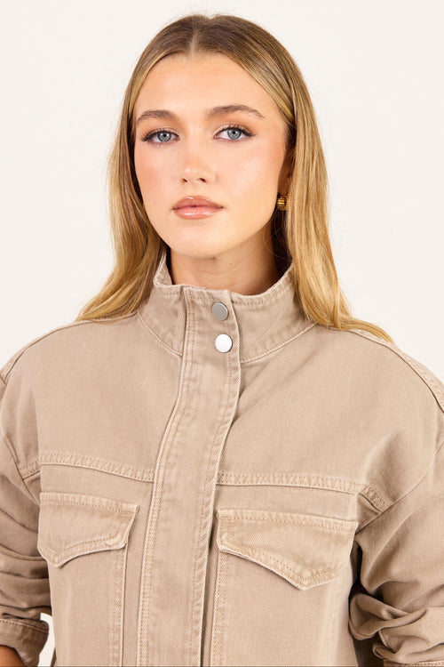 A blonde woman wearing a beige high-collar utility jacket with a snap-button placket and flap chest pockets, photographed against a clean off-white studio background.
