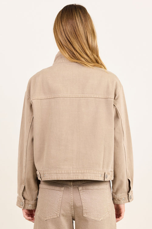 A woman viewed from behind wearing a cropped beige denim jacket and matching beige pants against a plain off-white studio background.