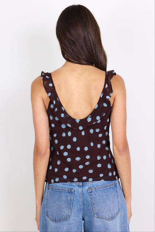 A woman viewed from the back wearing a dark brown sleeveless top with a light blue polka dot pattern and light blue denim jeans, set against a plain white background.