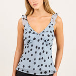 A woman modeling a light blue sleeveless top with black polka dots and ruffled shoulder details, paired with black pants, against a plain off-white background.