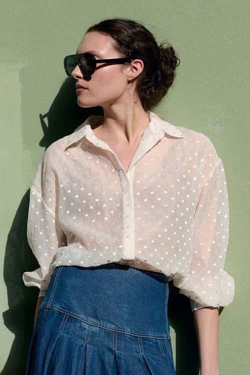 A woman stands against a light green wall wearing a sheer white polka-dot blouse, a long blue denim skirt, black loafers, and sunglasses, holding a dark brown handbag.