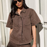 A woman wearing a matching brown short-sleeve button-up shirt and shorts set, styled with black sunglasses and a black handbag, standing in front of a white window with curtains.