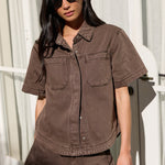 A woman wearing a matching brown short-sleeve button-up shirt and shorts set, styled with black sunglasses and holding a black handbag, standing in front of a white window frame.