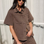 A woman wearing a matching brown short-sleeve button-up shirt and shorts set, styled with black sunglasses and holding a black handbag, posed in front of a white window with sheer curtains.