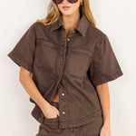 A woman modeling a matching brown short-sleeve button-up shirt and shorts set against a plain white background.