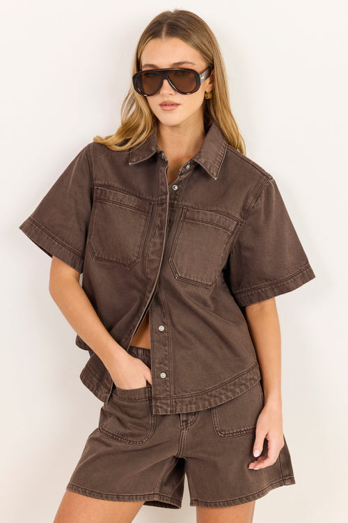A woman modeling a matching brown short-sleeve button-up shirt and shorts set against a plain white background.