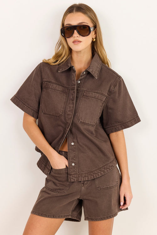 A woman modeling a matching brown short-sleeve button-up shirt and shorts set against a plain off-white background.
