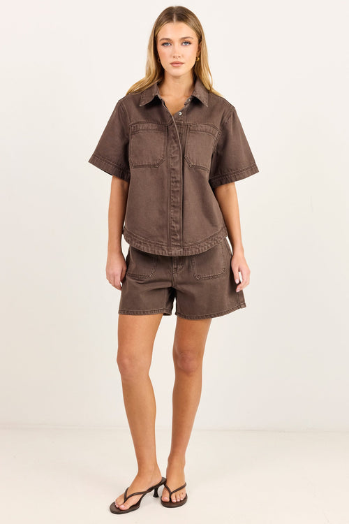 A woman modeling a matching brown short-sleeve button-up shirt and shorts set against a plain white studio background.