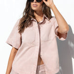A woman wearing a light pink short-sleeve button-up shirt and matching pink pants, styled with black sunglasses, posing against a bright white wall.