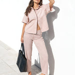 A woman stands by a white wall and poolside wearing a matching light blush pink short-sleeve button-up shirt and cropped trousers set. She is wearing black sunglasses, black sandals, and holding a black tote bag.