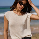 A woman wearing a cream-colored, short-sleeve knit top and black pants stands on a beach with the ocean and blue sky in the background.