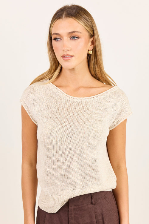 A woman modeling a short-sleeve knit top in a light cream or off-white color, paired with dark brown trousers, against a plain light background.