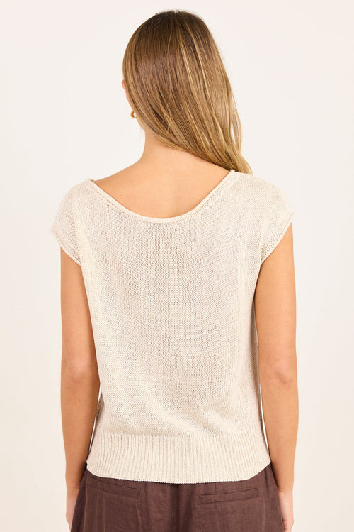 A woman viewed from the back wearing a light cream knit top with cap sleeves and a wide scoop neckline, paired with brown pants, against a plain off-white background.