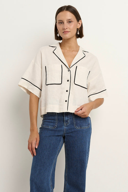 Woman wearing an oversized cream linen shirt with black piping, short sleeves, and front buttons, styled with blue denim jeans.