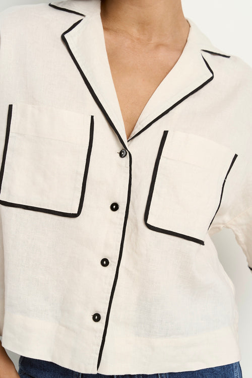 Woman wearing an oversized cream linen shirt with black piping, short sleeves, and front buttons, styled with blue denim jeans.