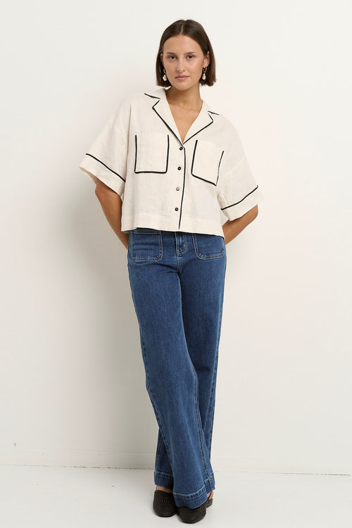 Woman wearing an oversized cream linen shirt with black piping, short sleeves, and front buttons, styled with blue denim jeans.