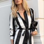 Woman wearing a black and white striped dress with sunglasses and clutch against a light-colored wall.