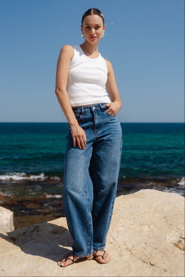 Woman standing on rocks by the ocean wearing a white top and blue jeans.