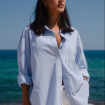 Woman standing on a beach wearing a light blue striped shirt and white pants.