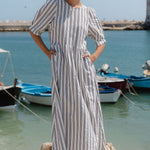 Woman in a striped dress standing on rocks by a harbor with boats and a clear sky.