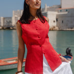 Woman in a red top and white skirt standing by a waterfront with buildings in the background.