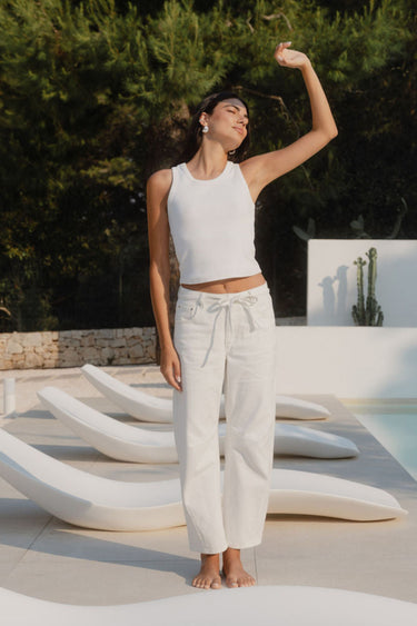 Woman in white outfit standing on a modern outdoor chair by a pool with greenery in the background