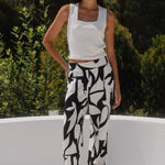 Woman wearing a white top and black and white patterned pants standing on a white architectural structure with greenery in the background.