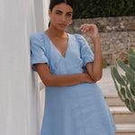 Woman in a light blue dress standing against a white wall with greenery in the background