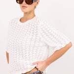 A woman modeling a white open-knit short-sleeve crop top with blue jeans and brown sunglasses against a plain white background.