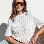 A woman wearing a white textured short-sleeve knit top and light blue jeans, sitting on a brown woven chair against a white wall.