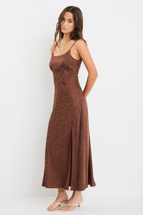 Woman wearing a chocolate brown strappy bias-cut midi dress with a subtle animal print and front slit, styled with cream heeled sandals.