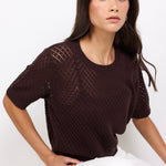 Woman wearing a dark brown knitted top and white pants against a white background