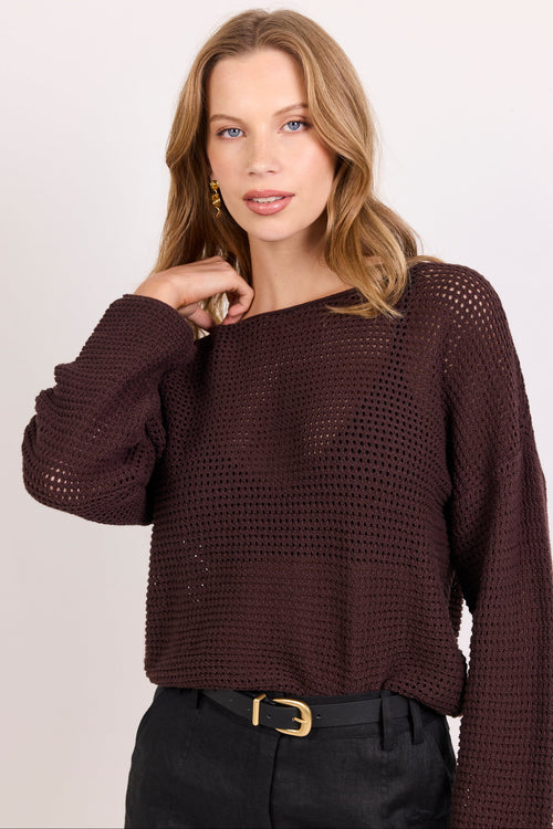 A woman wearing a dark brown open-knit long-sleeve sweater and black pants with a gold belt buckle, photographed against a light gray studio background.
