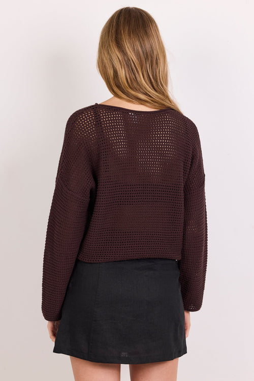 A woman shown from the back wearing a dark brown open-knit cropped sweater and a black mini skirt, against a plain light gray studio background.