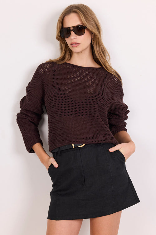 A woman modeling a dark brown textured knit sweater with a boat neckline and long sleeves, paired with a black mini skirt and sunglasses, against a plain white background.