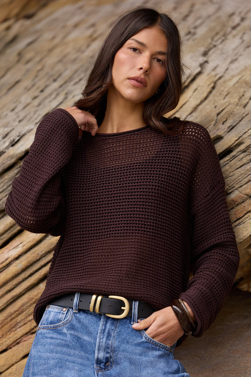 A woman wearing a dark brown open-knit long-sleeve sweater and blue jeans, posing against a textured rock background.
