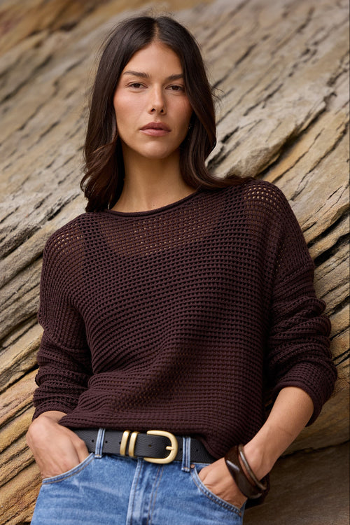 A woman stands outdoors against a large rock formation, wearing a dark brown open-knit long-sleeve sweater, blue denim shorts, and a black belt with gold hardware.