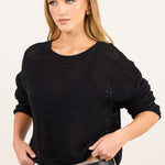 A woman modeling a black open-knit long-sleeve top with beige pants and a black belt, photographed against a plain light background.
