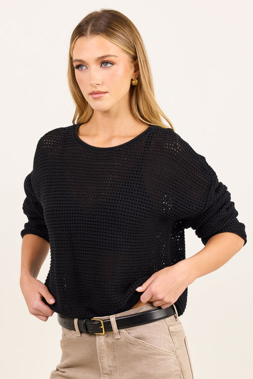 A woman modeling a black open-knit long-sleeve top with beige pants and a black belt, photographed against a plain light background.