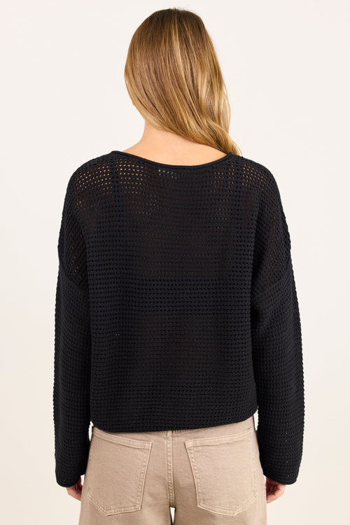 A woman viewed from the back wearing a black open-knit cropped sweater and beige pants against a plain off-white studio background.