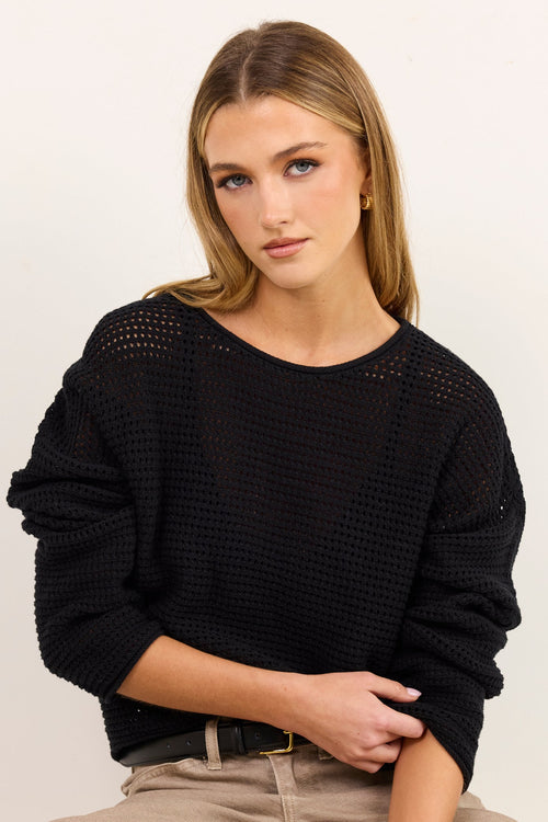 A woman wearing a black textured knit sweater and beige wide-leg pants, seated on a cream-colored stool against a plain off-white background.