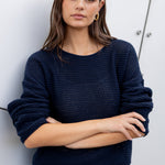 Woman wearing a navy sweater and blue jeans against a white wall.