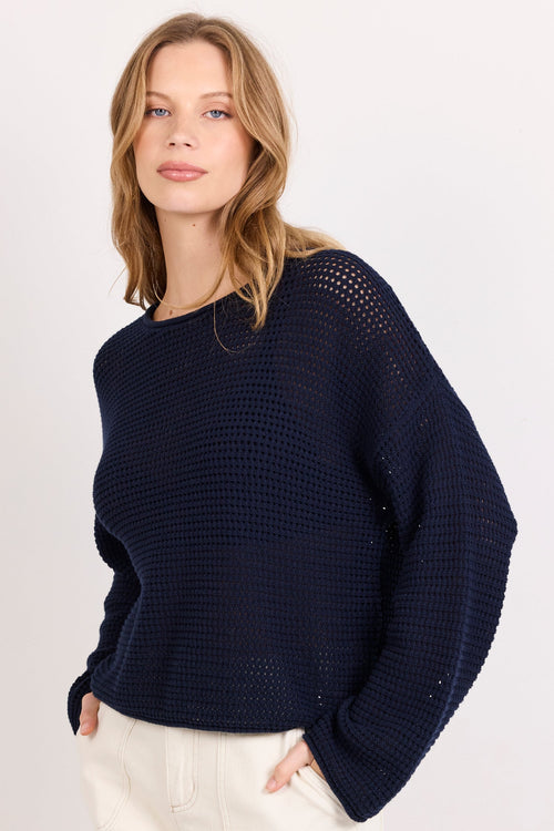 A woman modeling a dark navy blue knit sweater with textured openwork details on the shoulders, paired with cream shorts, against a plain light background.