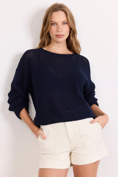 A woman modeling a dark navy blue knit sweater with textured openwork details and cream-colored shorts, standing against a plain white studio background.