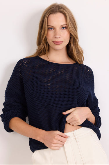 A woman wearing a dark navy blue open-knit long-sleeve sweater and off-white pants, standing against a plain white background.